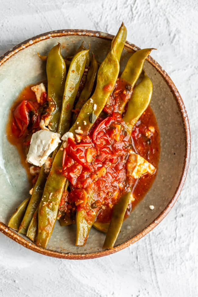 Eine portion geschmorte Stangenbohnen in Tomatensauce mit Feta auf Keramikteller und weißem Untergrund.