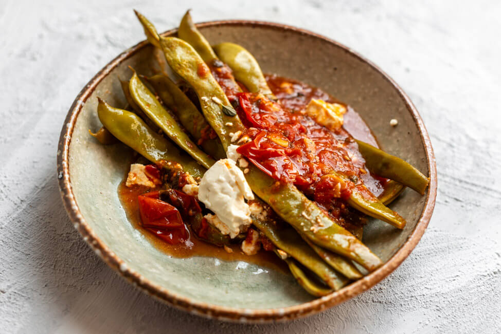 Eine portion geschmorte Stangenbohnen in Tomatensauce mit Feta auf Keramikteller und weißem Untergrund.