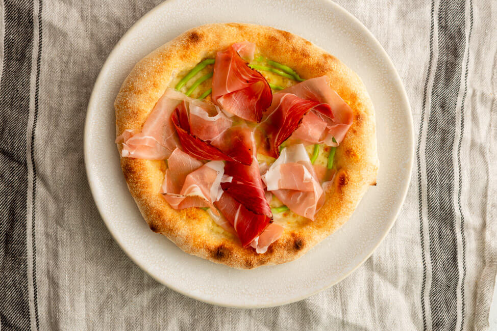 Pizza mit Südtiroler Speck und Schnittlauchmayonnaise auf weißem Pizzateller.