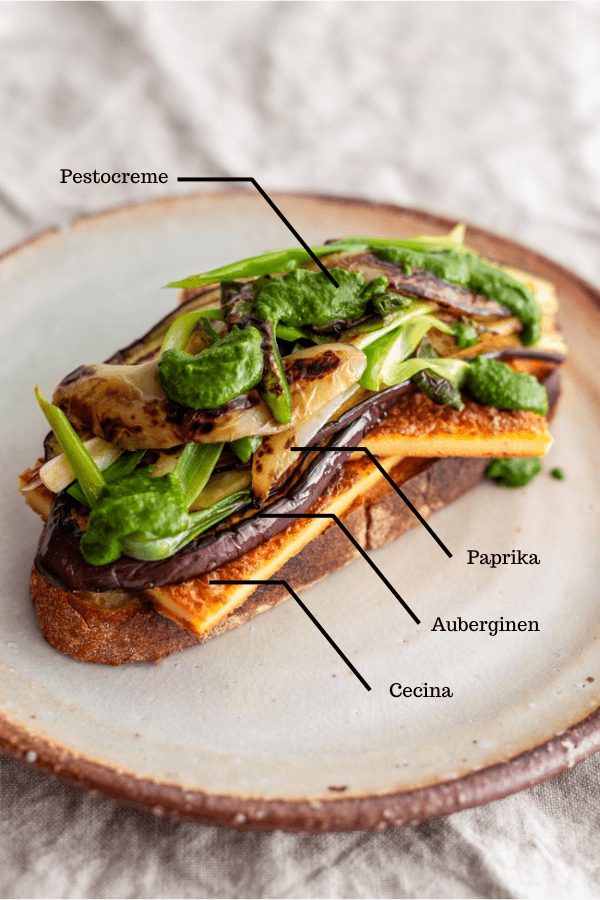 Belegte Scheibe italienisches Weißbrot mit Cecina, Auberginen und Paprika Antipasti und Pestocreme auf Keramikteller auf hellem Tischtuch