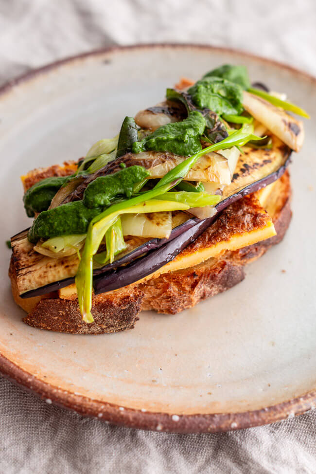 Belegte Scheibe italienisches Weißbrot mit Cecina, Auberginen und Paprika Antipasti und Pestocreme auf Keramikteller auf hellem Tischtuch
