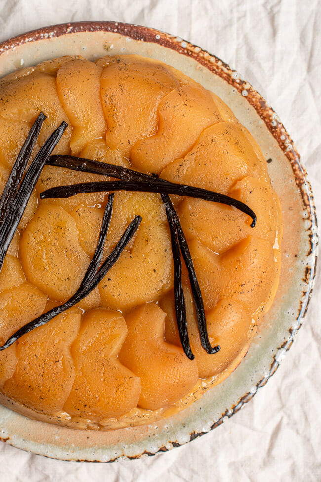 Tarte Tatin mit Vanilleschoten dekoriert auf Kuchenplatte und heller Tischdecke.