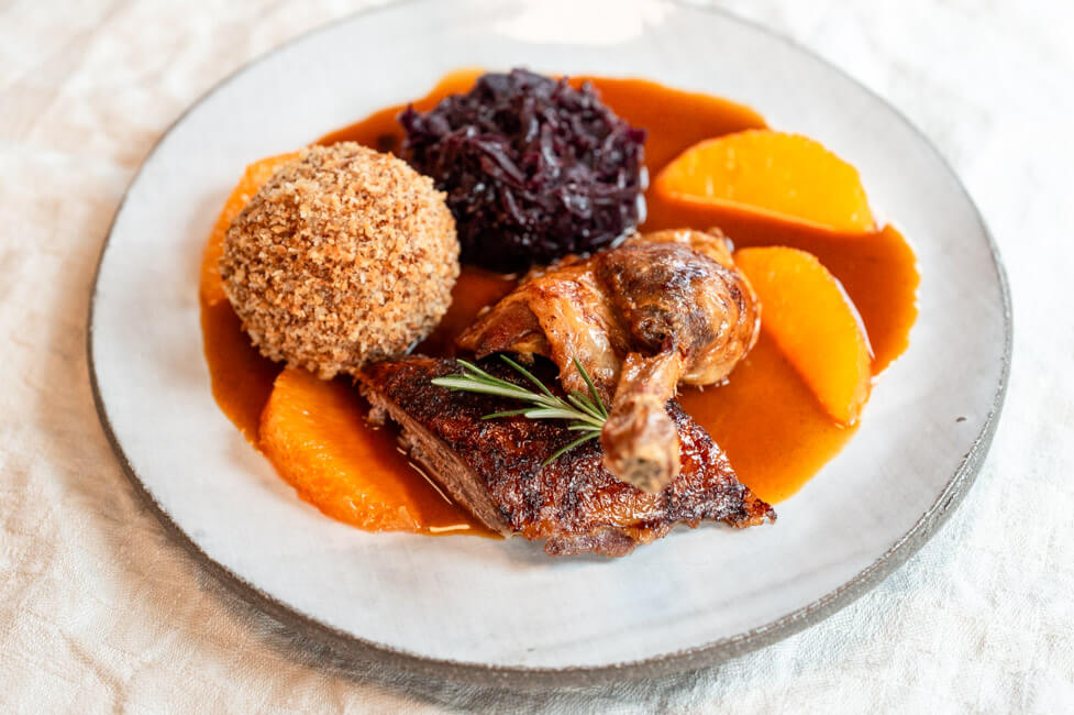 Bauernernte mit Klößen, Rotkohl und Orangenjus aus Kitchen Impossible auf hellem Teller und cremefarbener Tischdecke.