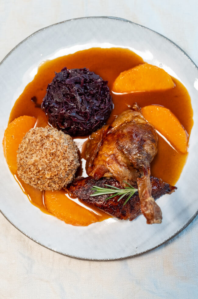 Bauernernte mit Klößen, Rotkohl und Orangenjus aus Kitchen Impossible auf hellem Teller und cremefarbener Tischdecke.