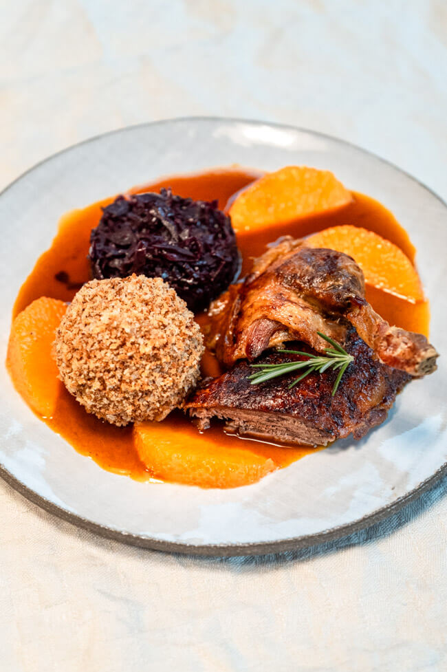 Bauernernte mit Klößen, Rotkohl und Orangenjus aus Kitchen Impossible auf hellem Teller und cremefarbener Tischdecke.
