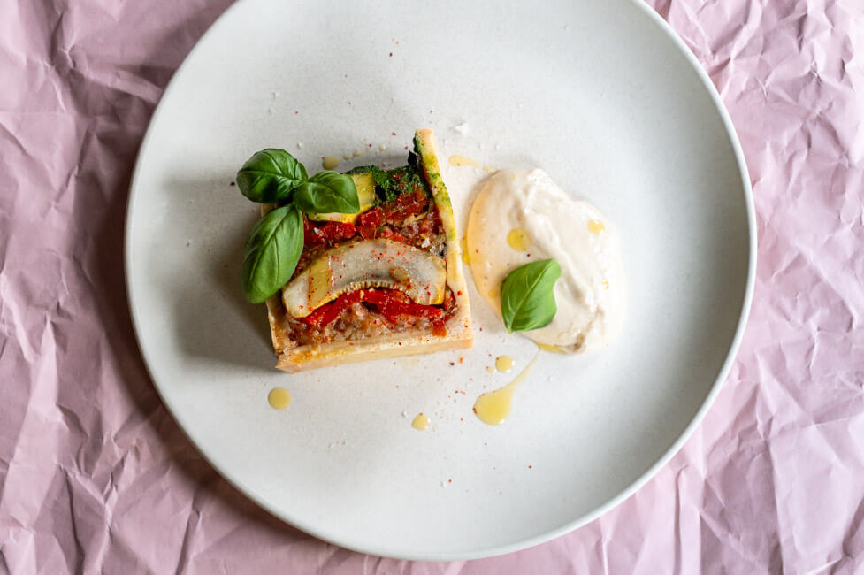 Vegetarische Pâté en croûte mit Basilikum Coulis und Anchovis-Soße angerichtet auf hellem Teller mit Olivenöl, grobem Salt und Piment d'Espelett und Basilikumblättern garniert.