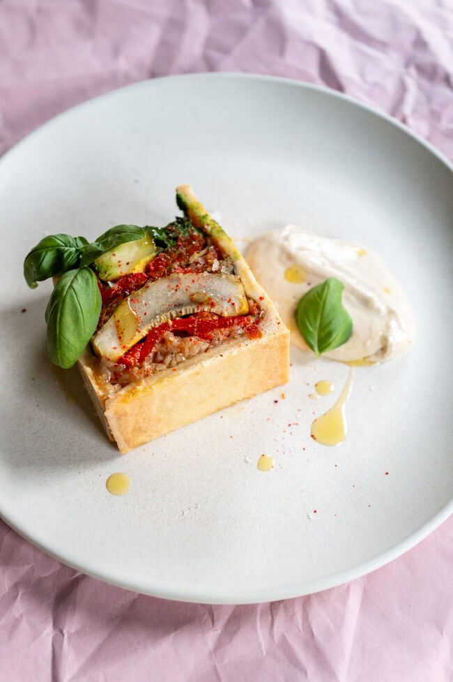 Vegetarische Pâté en croûte mit Basilikum Coulis und Anchovis-Soße angerichtet auf hellem Teller mit Olivenöl, grobem Salt und Piment d'Espelett und Basilikumblättern garniert.