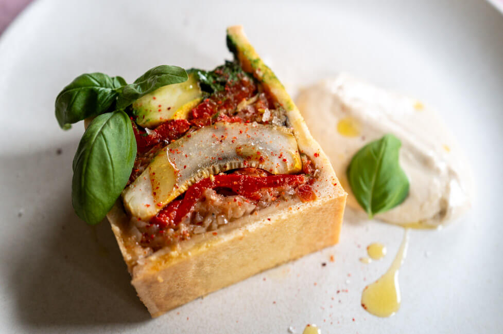 Vegetarische Pâté en croûte mit Basilikum Coulis und Anchovis-Soße angerichtet auf hellem Teller mit Olivenöl, grobem Salt und Piment d'Espelett und Basilikumblättern garniert.