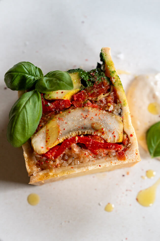 Vegetarische Pâté en croûte mit Basilikum Coulis und Anchovis-Soße angerichtet auf hellem Teller mit Olivenöl, grobem Salt und Piment d'Espelett und Basilikumblättern garniert.