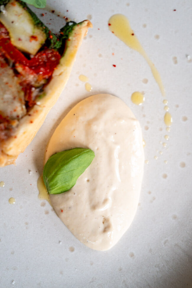 Vegetarische Pâté en croûte mit Basilikum Coulis und Anchovis-Soße angerichtet auf hellem Teller mit Olivenöl, grobem Salt und Piment d'Espelett und Basilikumblättern garniert.