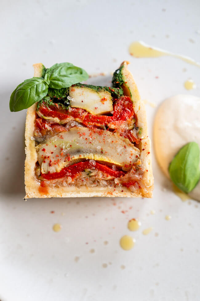 Vegetarische Pâté en croûte mit Basilikum Coulis und Anchovis-Soße angerichtet auf hellem Teller mit Olivenöl, grobem Salt und Piment d'Espelett und Basilikumblättern garniert.