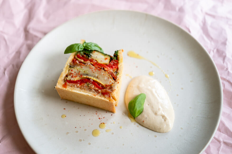 Vegetarische Pâté en croûte mit Basilikum Coulis und Anchovis-Soße angerichtet auf hellem Teller mit Olivenöl, grobem Salt und Piment d'Espelett und Basilikumblättern garniert.