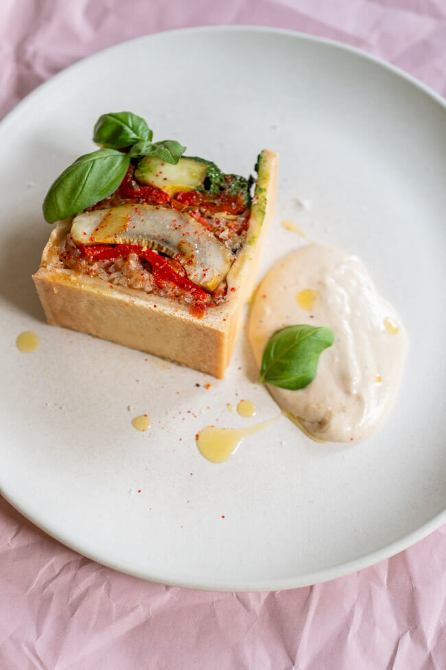 Vegetarische Pâté en croûte mit Basilikum Coulis und Anchovis-Soße angerichtet auf hellem Teller mit Olivenöl, grobem Salt und Piment d'Espelett und Basilikumblättern garniert.