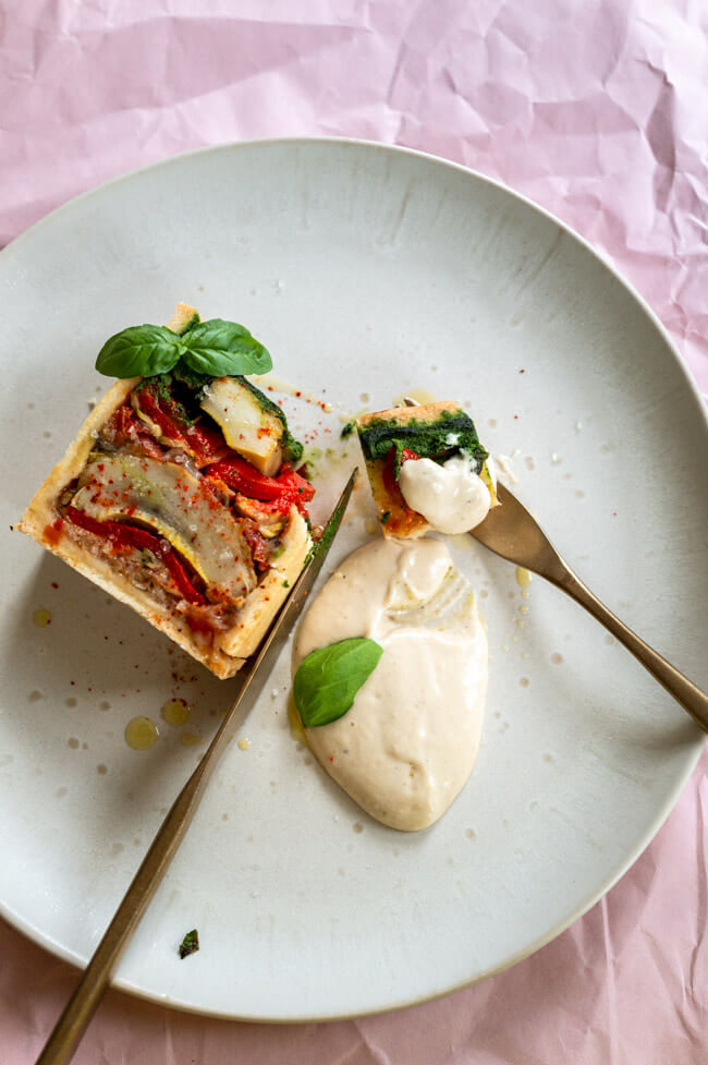 Vegetarische Pâté en croûte mit Basilikum Coulis und Anchovis-Soße angerichtet auf hellem Teller mit Olivenöl, grobem Salt und Piment d'Espelett und Basilikumblättern garniert.