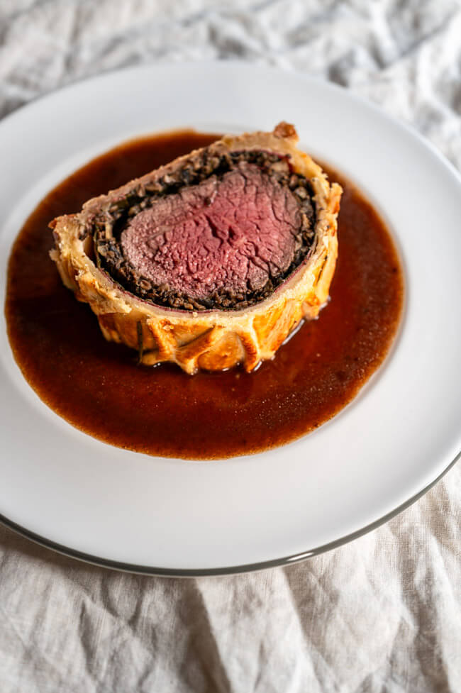 Eine Scheibe Beef Wellington mit Jus auf hellem Porzellanteller und heller Tischdecke