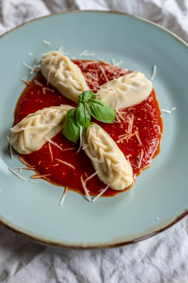 4 Culurgiones mit Tomatensauce, Parmesan und frischem Basilikum auf blauem Teller angerichtet