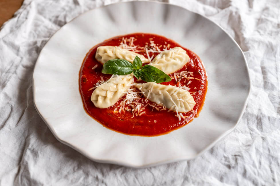 4 Culurgiones mit Tomatensauce, Parmesan und frischem Basilikum auf hellem Teller angerichtet