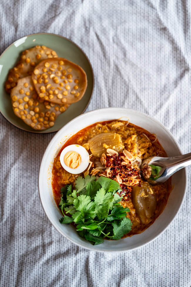 Eine Schüssel Mohinga, getoppt mit Koriander, Ei, Chili und Knoblauch dazu ein Teller mit Chana Dal Cracker auf heller Tischdecke.