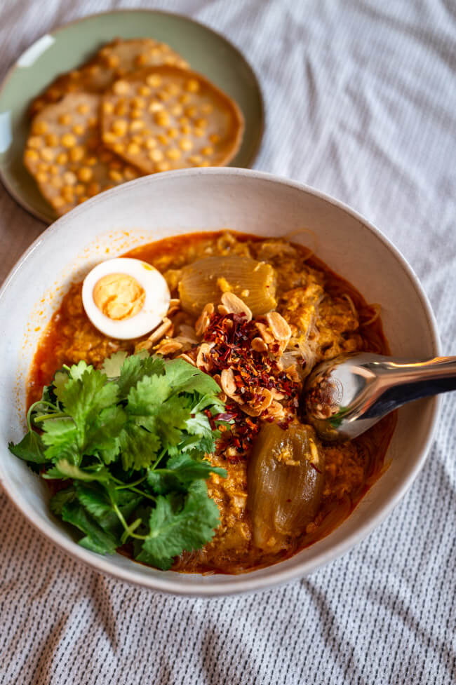 Eine Schüssel Mohinga, getoppt mit Koriander, Ei, Chili und Knoblauch dazu ein Teller mit Chana Dal Cracker auf heller Tischdecke.