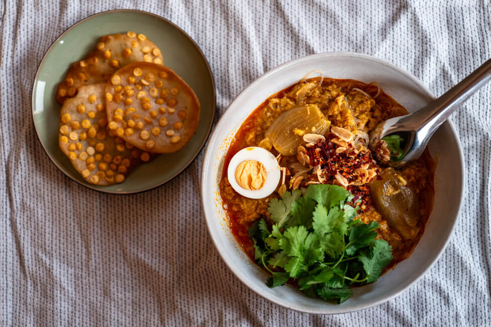 Eine Schüssel Mohinga, getoppt mit Koriander, Ei, Chili und Knoblauch dazu ein Teller mit Chana Dal Cracker auf heller Tischdecke.