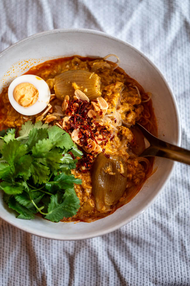 Eine Schüssel Mohinga, getoppt mit Koriander, Ei, Chili und Knoblauch auf heller Tischdecke.