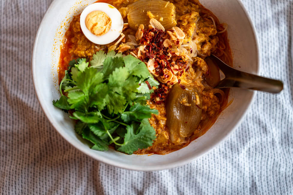 Eine Schüssel Mohinga, getoppt mit Koriander, Ei, Chili und Knoblauch auf heller Tischdecke.