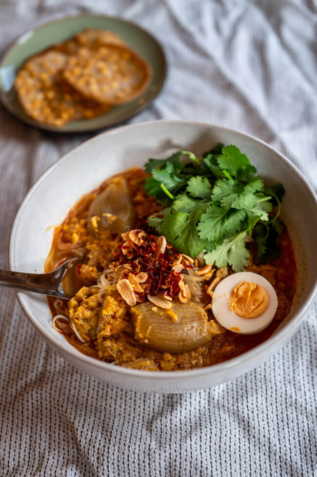 Eine Schüssel Mohinga, getoppt mit Koriander, Ei, Chili und Knoblauch dazu ein Teller mit Chana Dal Cracker auf heller Tischdecke.