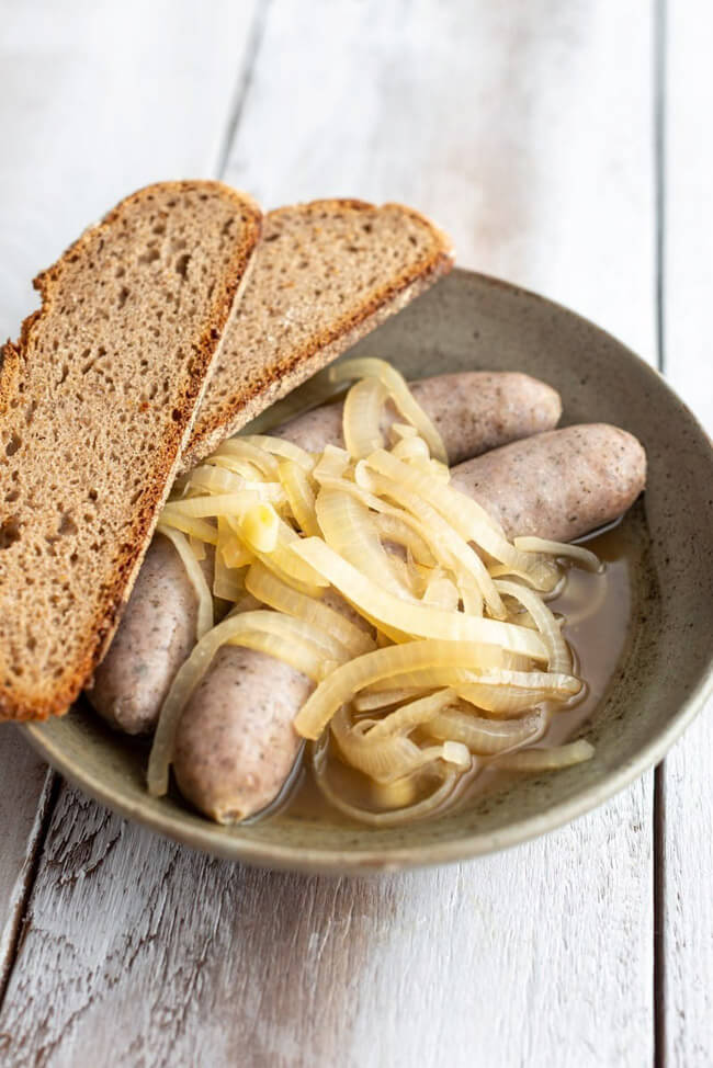 Bratwürste mit Zwiebeln in Essigsud liegen in grauem Teller mit zwei Scheiben Bauernbrot auf hellem Holztisch.