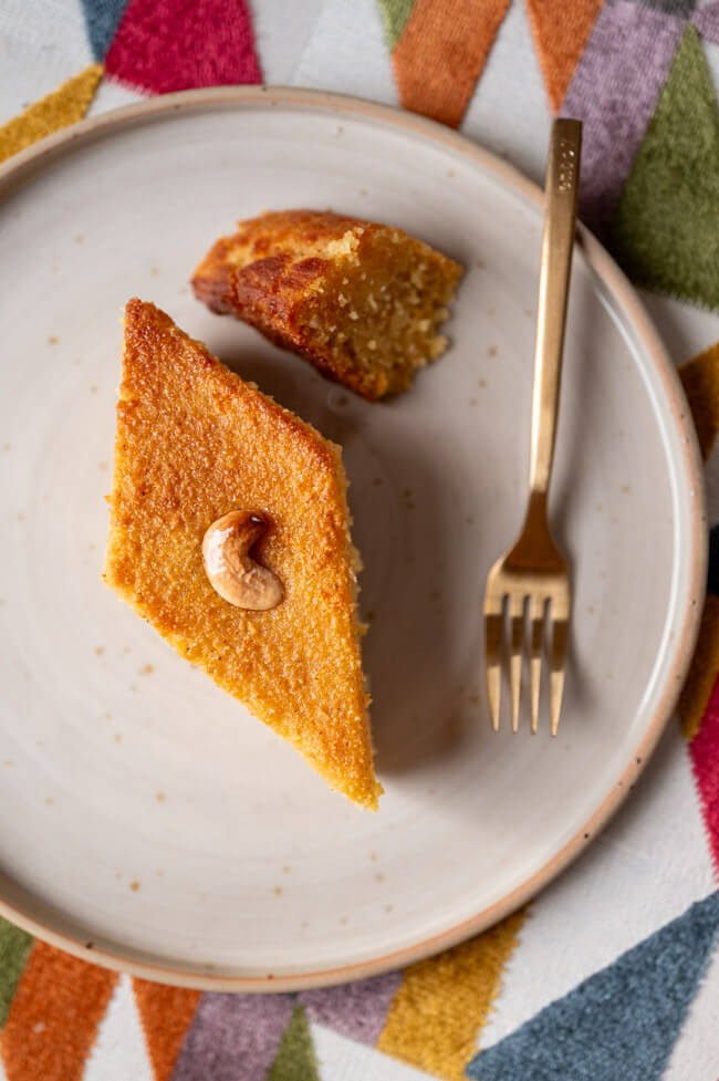Ein Stück Orangen-Grießkuchen mit Cashewkern belegt und goldener Gabel auf hellem Keramikteller