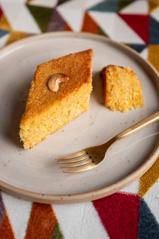 Ein Stück Orangen-Grießkuchen mit Cashewkern belegt und goldener Gabel auf hellem Keramikteller
