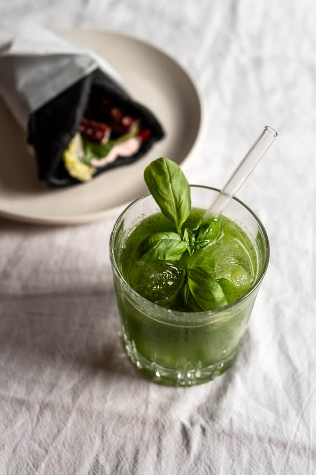 Gin Basil Smash angerichtet in Tumbler mit Eis und Baislikumblatt und Sepia-Pita mit Oktopus im Hintergrund