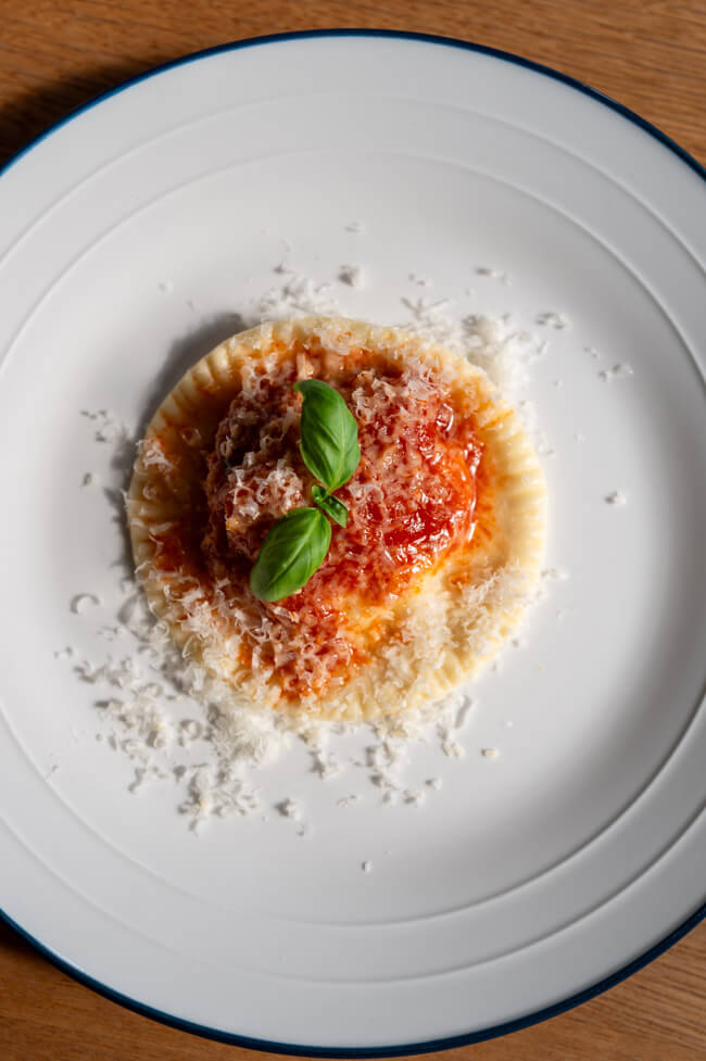 Ravioli Gallurese angerichtet auf weißem Teller mit Tomatensoße, geriebenem Parmesan und Basilikum