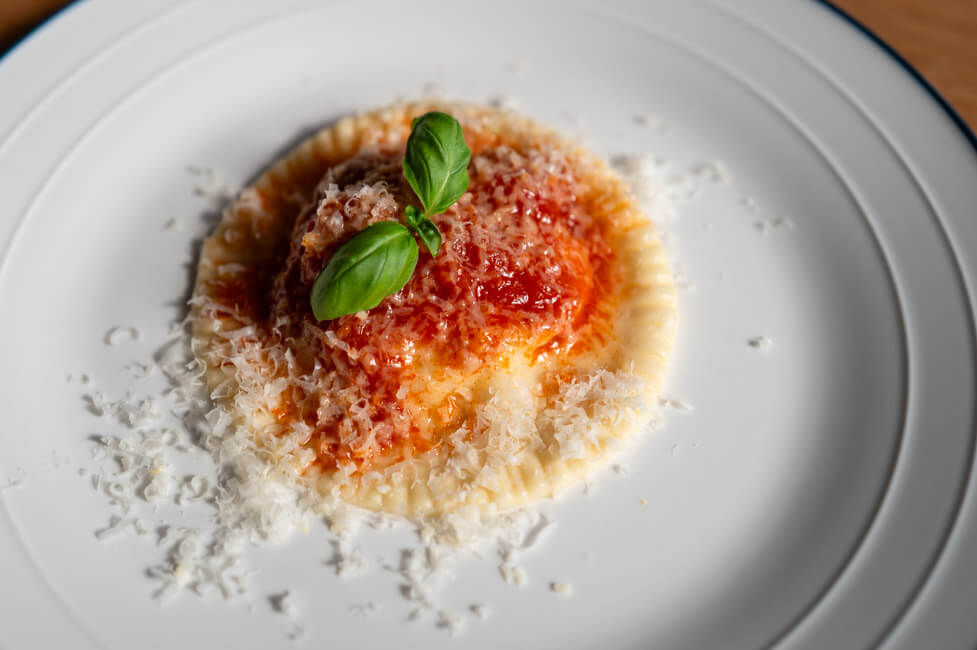 Ravioli Gallurese angerichtet auf weißem Teller mit Tomatensoße, geriebenem Parmesan und Basilikum