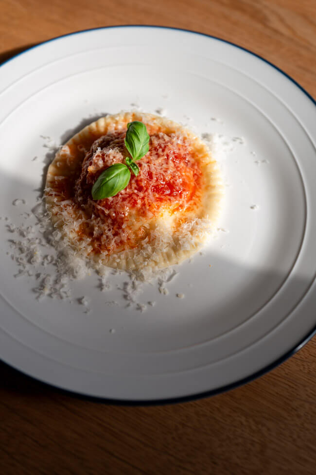 Ravioli Gallurese angerichtet auf weißem Teller mit Tomatensoße, geriebenem Parmesan und Basilikum