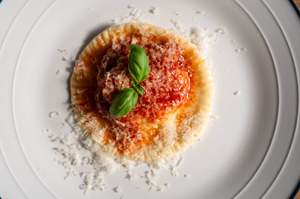 Ravioli Gallurese angerichtet auf weißem Teller mit Tomatensoße, geriebenem Parmesan und Basilikum