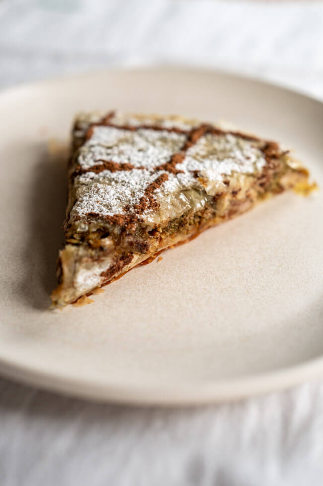 Ein Stück Pastilla, verziert mit Puderzucker und Zimt, auf Keramikteller und weißer Tischdecke