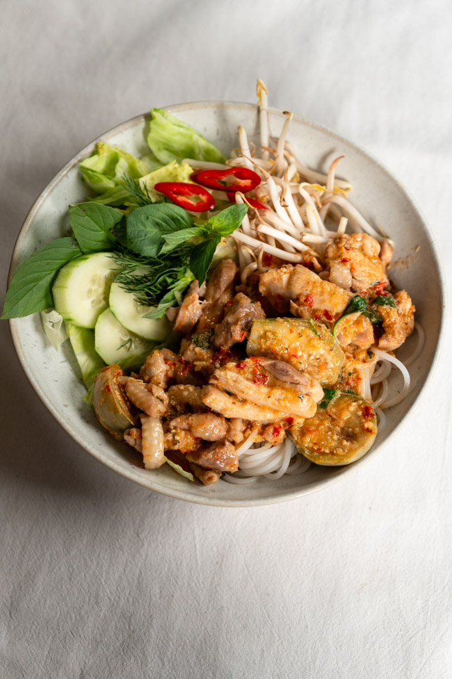 Eine Portion Gang Gai Korat mit Thai-Auberginen, frischen Sprossen, Thai Basilikum und Reisnudeln angerichtet auf weißem Teller