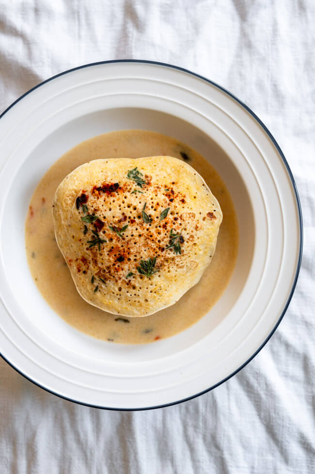 Eine Kaninchen-Frikadelle mit Polenta-Pfannkuchen mit Thymian und Pul Biber und Rahmsoße mit Meerrettich, Karotten und Sauerkrautsaft in weißem Teller mit blauem Rand