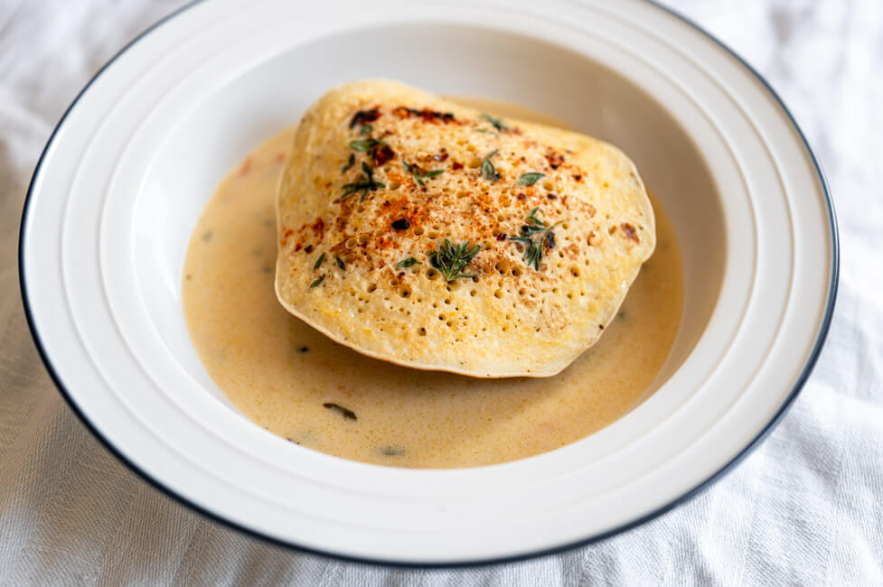 Eine Kaninchen-Frikadelle mit Polenta-Pfannkuchen mit Thymian und Pul Biber und Rahmsoße mit Meerrettich, Karotten und Sauerkrautsaft in weißem Teller mit blauem Rand