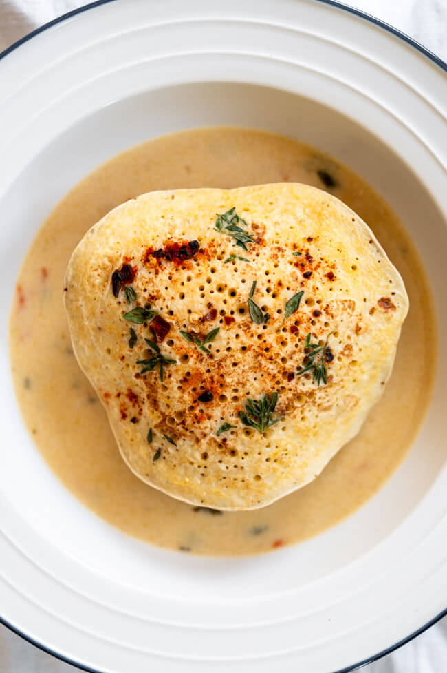 Eine Kaninchen-Frikadelle mit Polenta-Pfannkuchen mit Thymian und Pul Biber und Rahmsoße mit Meerrettich, Karotten und Sauerkrautsaft in weißem Teller mit blauem Rand
