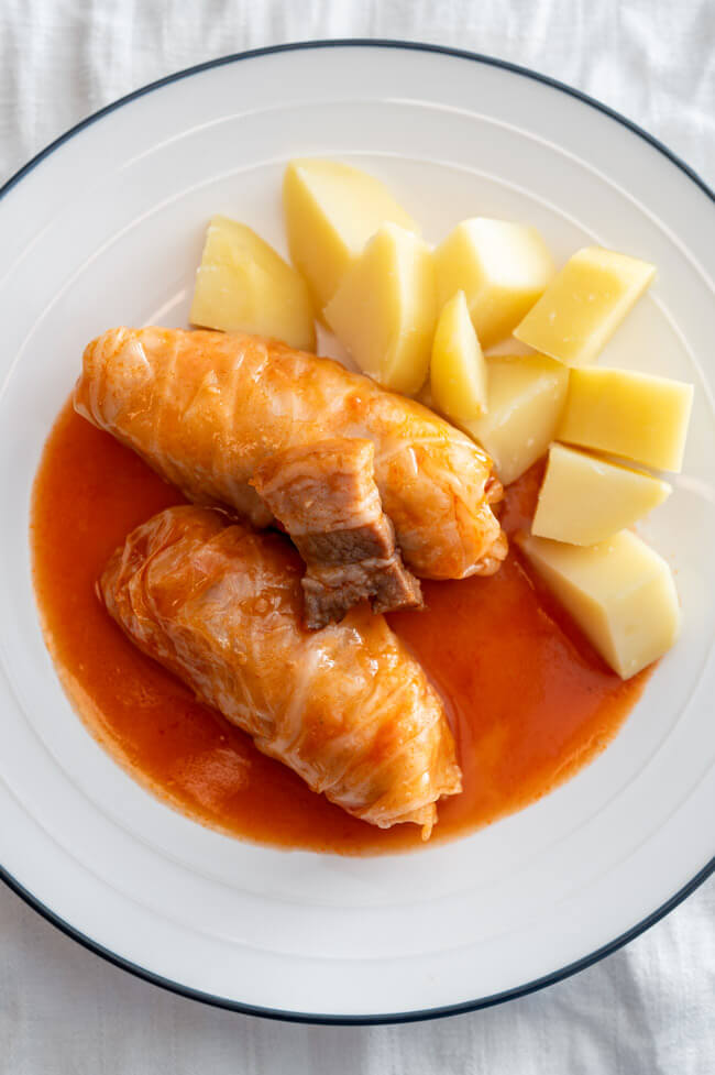 Zwei Krautwickel mit Tomatensoße und gekochten Kartoffeln und einem Stück Speck auf weißem Teller angerichtet