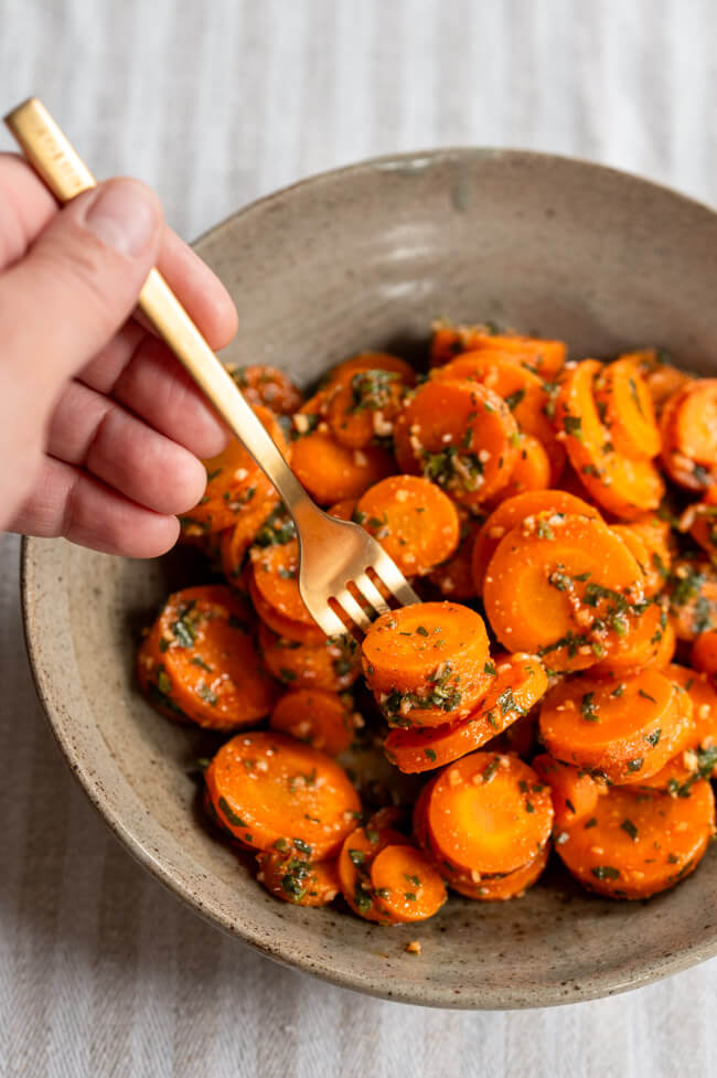 Hand hält Gabel mit Marokkanischem Karottensalat mit Koriander und Knoblauch in grauem Keramikteller auf hellem Untergrund