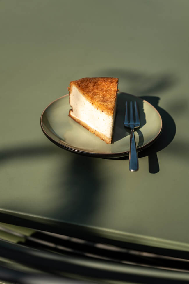 Ein Stück Haselnuss-Käsekuchen auf grünem Teller mit Gabel auf grünem Tisch im Sonnenlicht