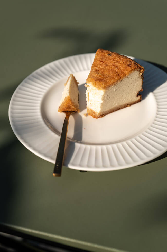 Ein Stück Haselnuss-Käsekuchen auf weißem Teller mit Gabel auf grünem Tisch im Sonnenlicht